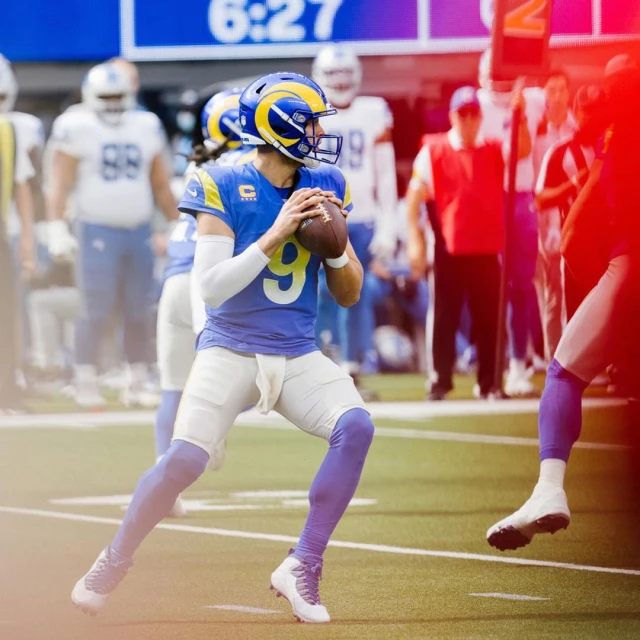 Football player in action on field, wearing team uniform with number 9, holding the football, stadium seating in background.