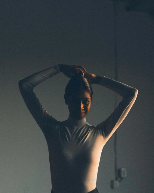 Woman in a white turtleneck bodysuit poses indoors, silhouetted against a dark background in a studio setting with rim lighting.