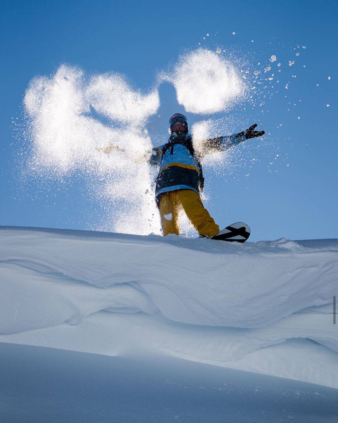 A snowboarder pauses on a snowdrift, captured from a low angle against a clear blue sky, with a burst of snow around the figure.
