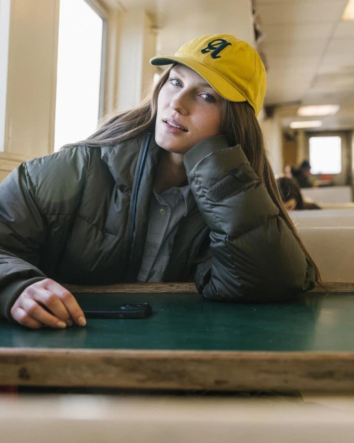 A woman with long hair sits at a table wearing a yellow cap and a green puffer jacket indoors, looking at the camera.