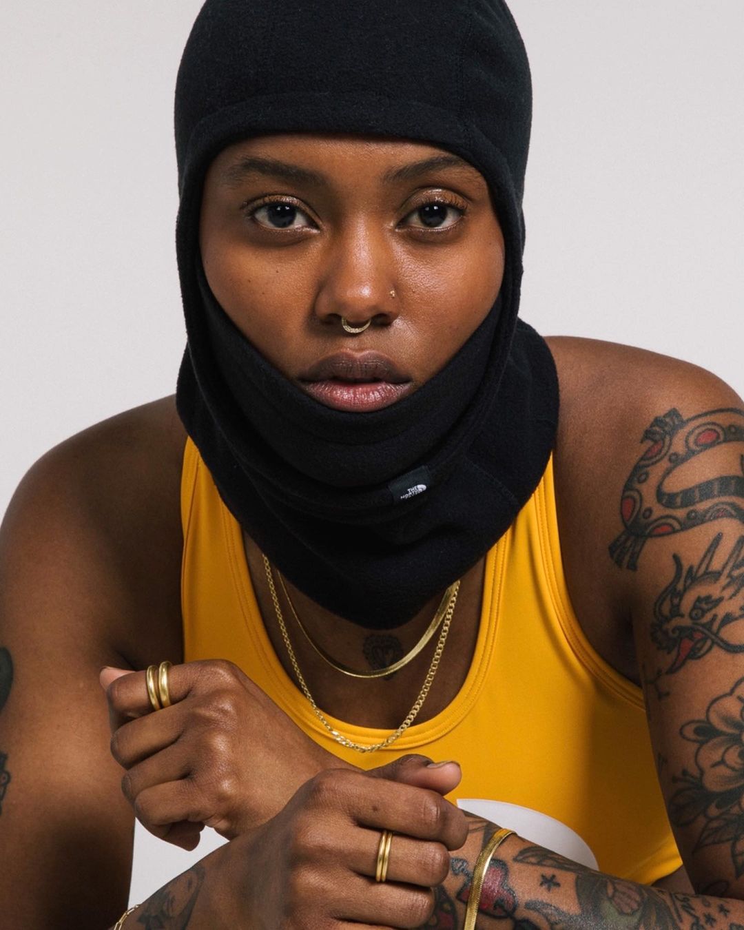 Close-up shot of a woman with dark skin wearing a black balaclava, yellow tank top, and gold jewelry, posing against a neutral background.