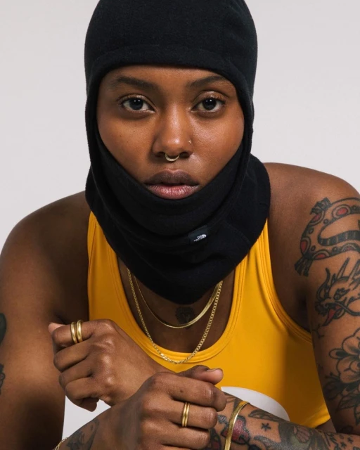Close-up shot of a woman with dark skin wearing a black balaclava, yellow tank top, and gold jewelry, posing against a neutral background.