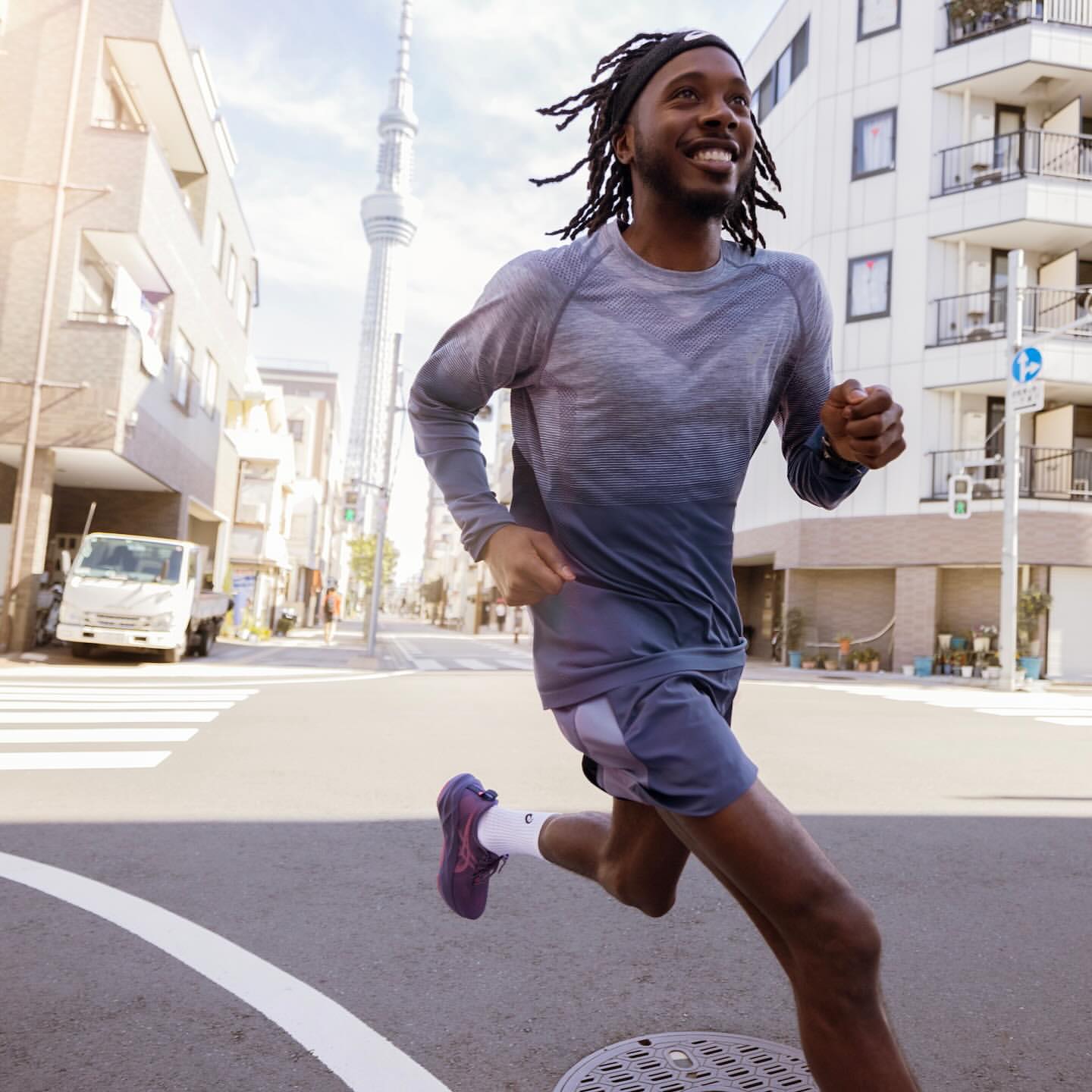 Athletic African-American man runs along a city street in workout gear with city buildings and tower in the background on a bright day.