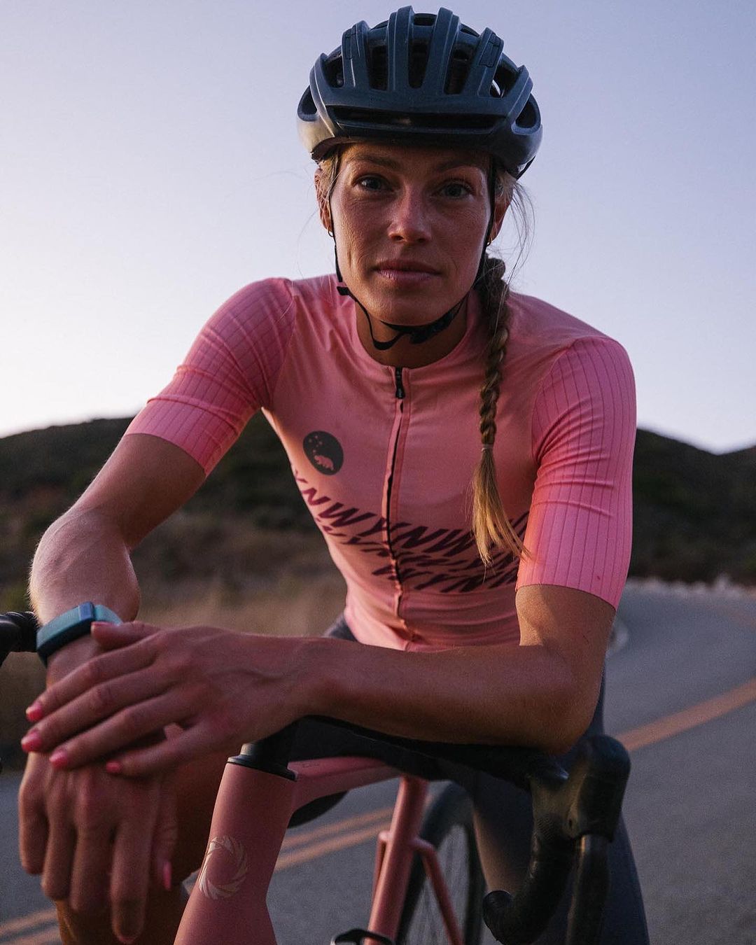 Woman in pink cycling jersey and helmet leaning on road bike on asphalt road in the evening.