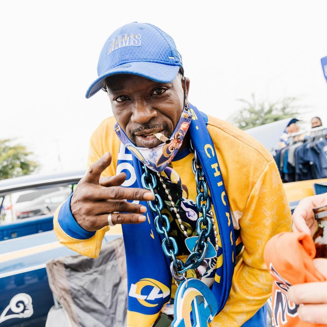Man smoking outdoors wearing a blue Rams cap and a gold and blue Rams shirt and necklace, flashing a hand sign with his fingers.
