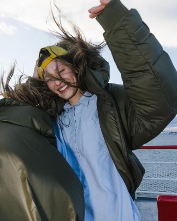 Smiling woman in an olive green puffer jacket and blue shirt with wind-blown hair outdoors on a boat.