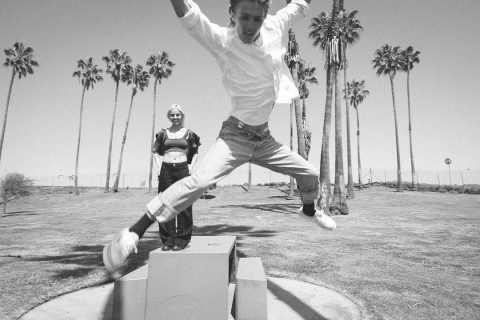 Black and white shot of a young man jumping off concrete blocks while a young woman stands nearby in a park with palm trees.