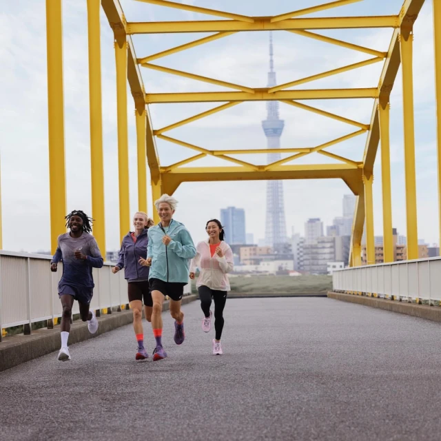 Four diverse athletes jog across a yellow bridge, Tokyo tower and city skyline in distance, fitness promotion, outdoor workout.