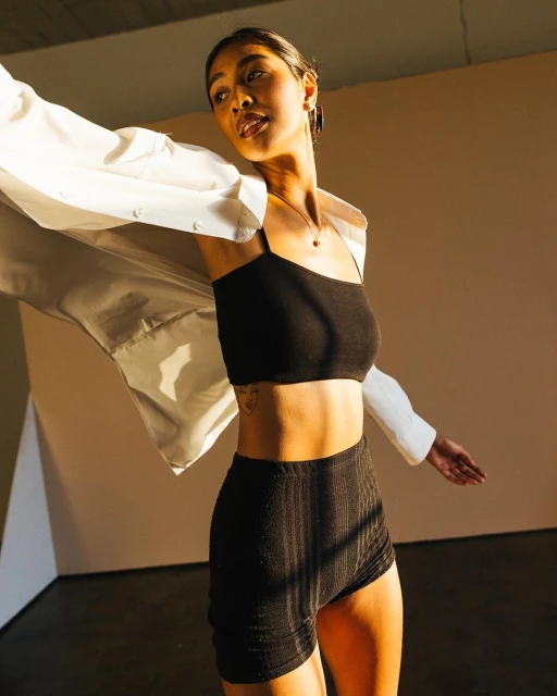 Stylish woman in black top and shorts with white shirt, posing in studio with warm lighting and dark floor. Fashion photography.