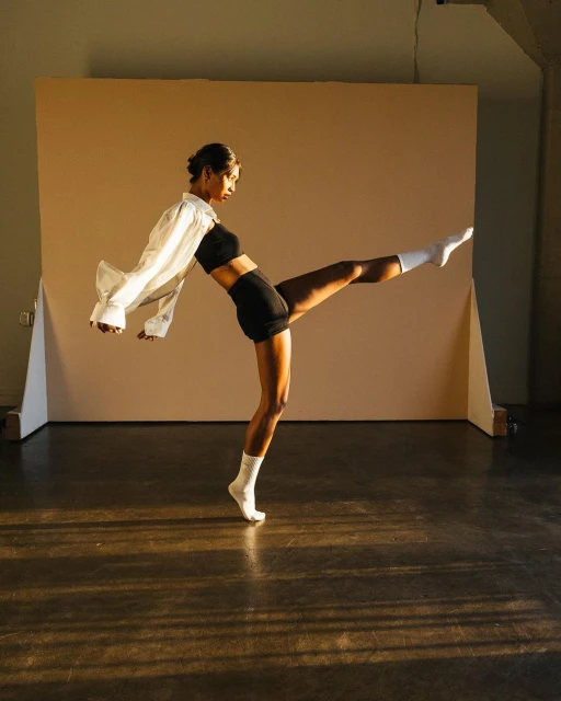 Athletic woman in black shorts and crop top, white socks and open shirt performing dynamic pose in studio with neutral background.