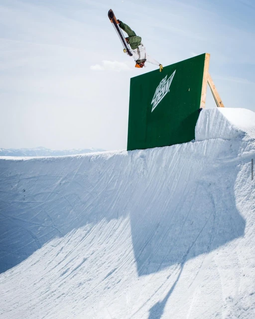 Snowboarder performing a trick off a green ramp with the Mountain Dew logo, set against a clear blue sky in a winter landscape.