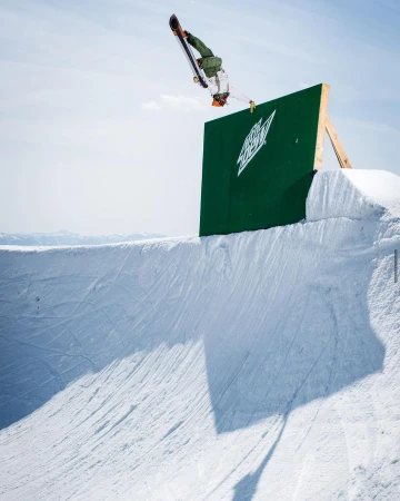 Snowboarder performing a trick off a green ramp with the Mountain Dew logo, set against a clear blue sky in a winter landscape.
