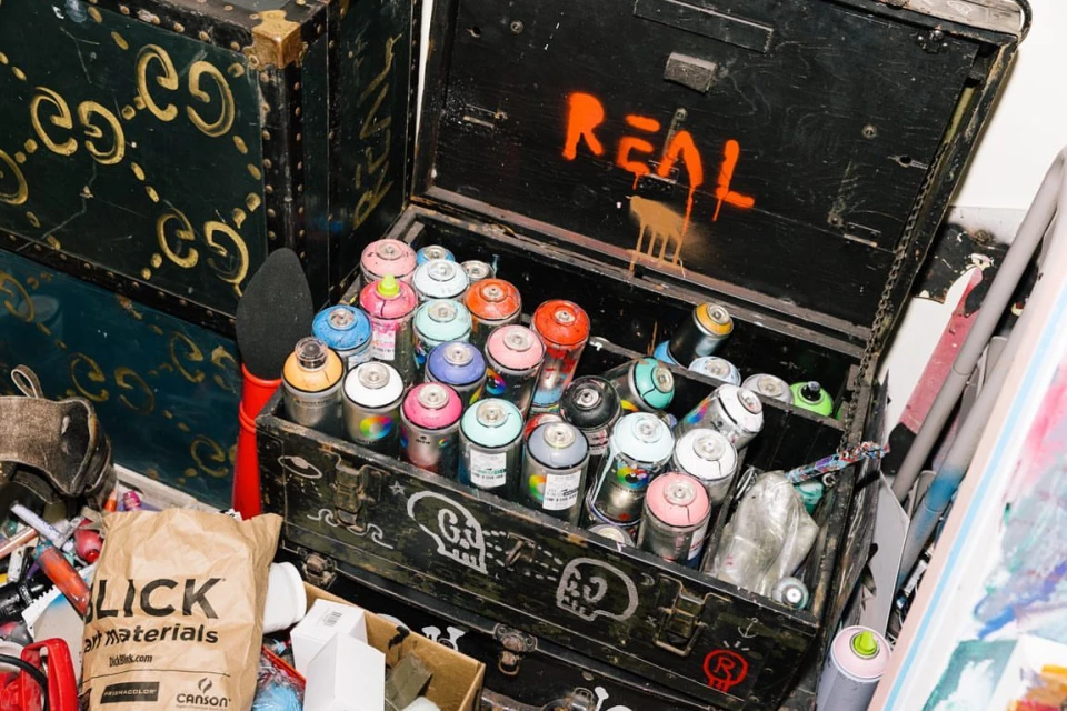 An open, black metal trunk filled with colorful spray paint cans sits next to a green Gucci trunk; art supplies and grunge aesthetic.