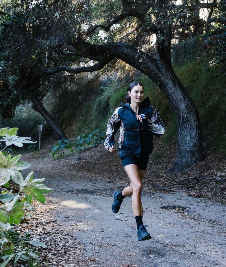 Athletic adult female runs on dirt path, wearing black shorts and a camouflage jacket, with trees and greenery in the background.