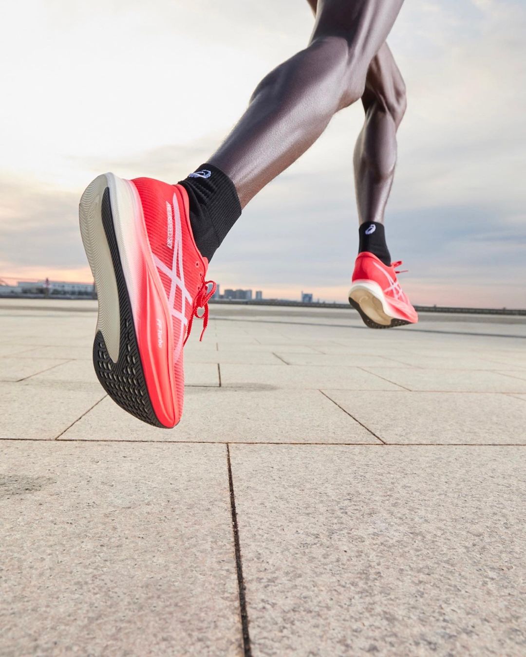 Close-up of a runner wearing bright red running shoes with visible ASICS logo, captured in motion on a concrete surface.