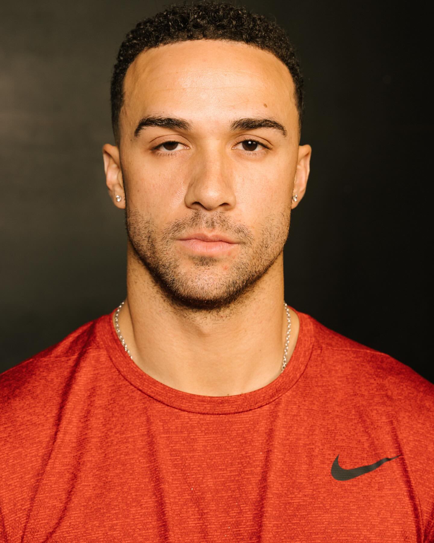 Portrait of a young adult man with brown hair wearing a red t-shirt with a black logo against a dark background, head and shoulders shot.