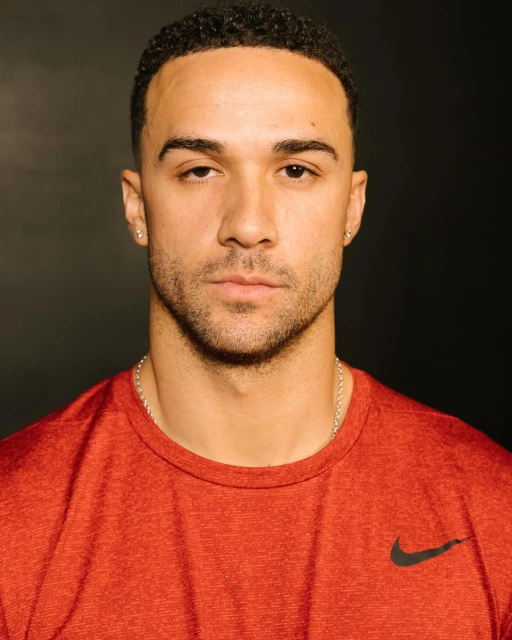 Portrait of a young adult man with brown hair wearing a red t-shirt with a black logo against a dark background, head and shoulders shot.