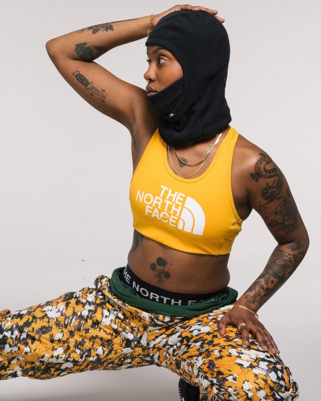 Woman in yellow sports bra and black balaclava strikes a dynamic pose in front of a solid white backdrop, showcasing a modern, sporty aesthetic.