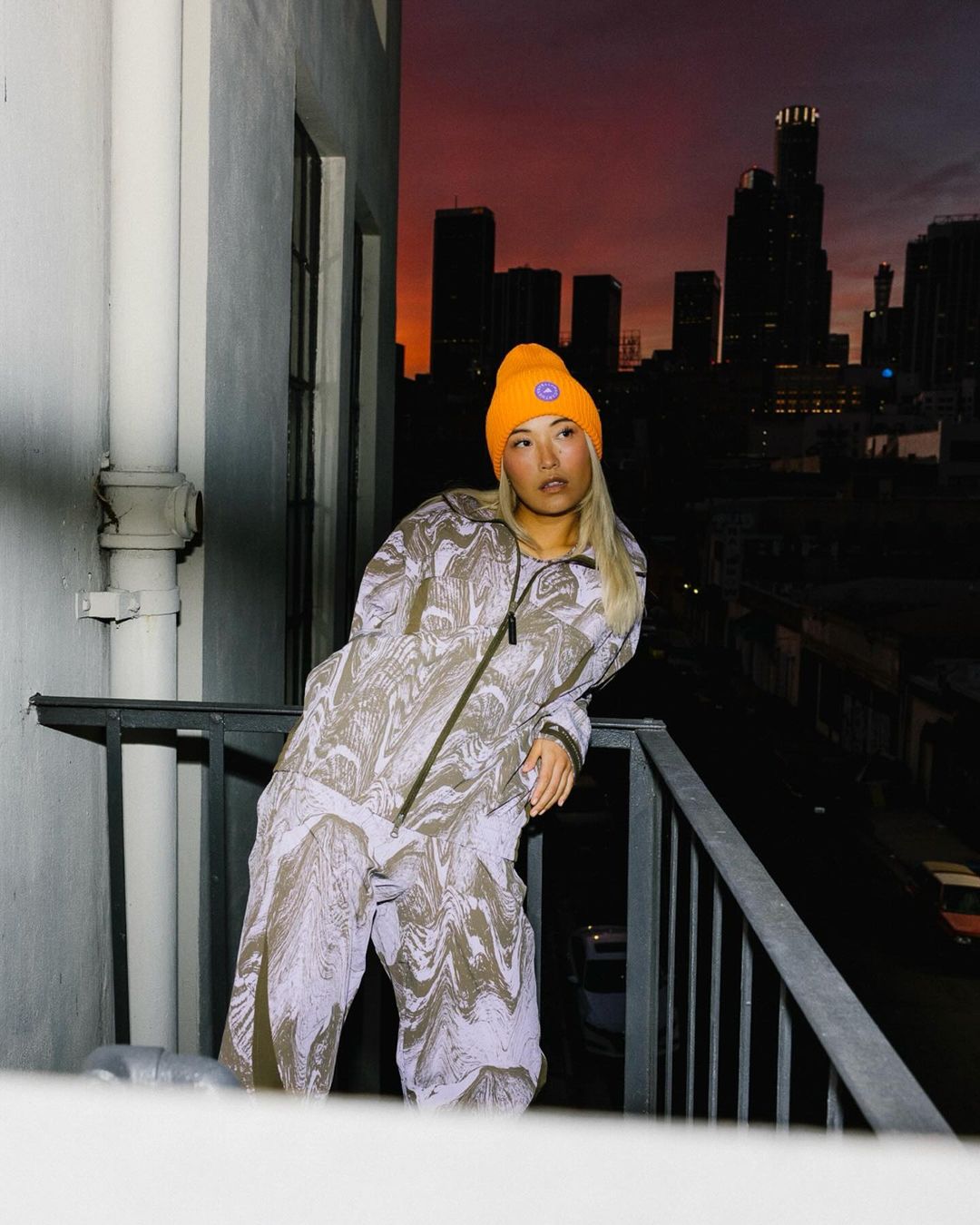 Young Asian woman poses on balcony in patterned jumpsuit and bright orange beanie against a dusk cityscape backdrop, fashion-forward.
