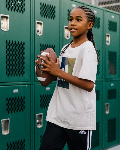 A young Black child holds an American football in front of a green locker backdrop, wearing casual clothing and braids.