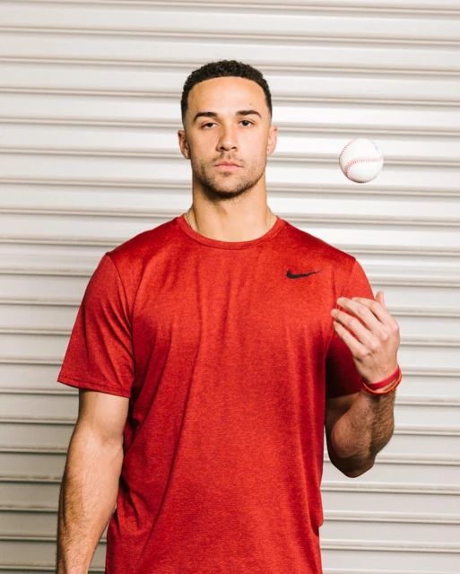 Athletic male baseball player in red Nike tee shirt tossing a baseball in a photo studio with a roller door background.
