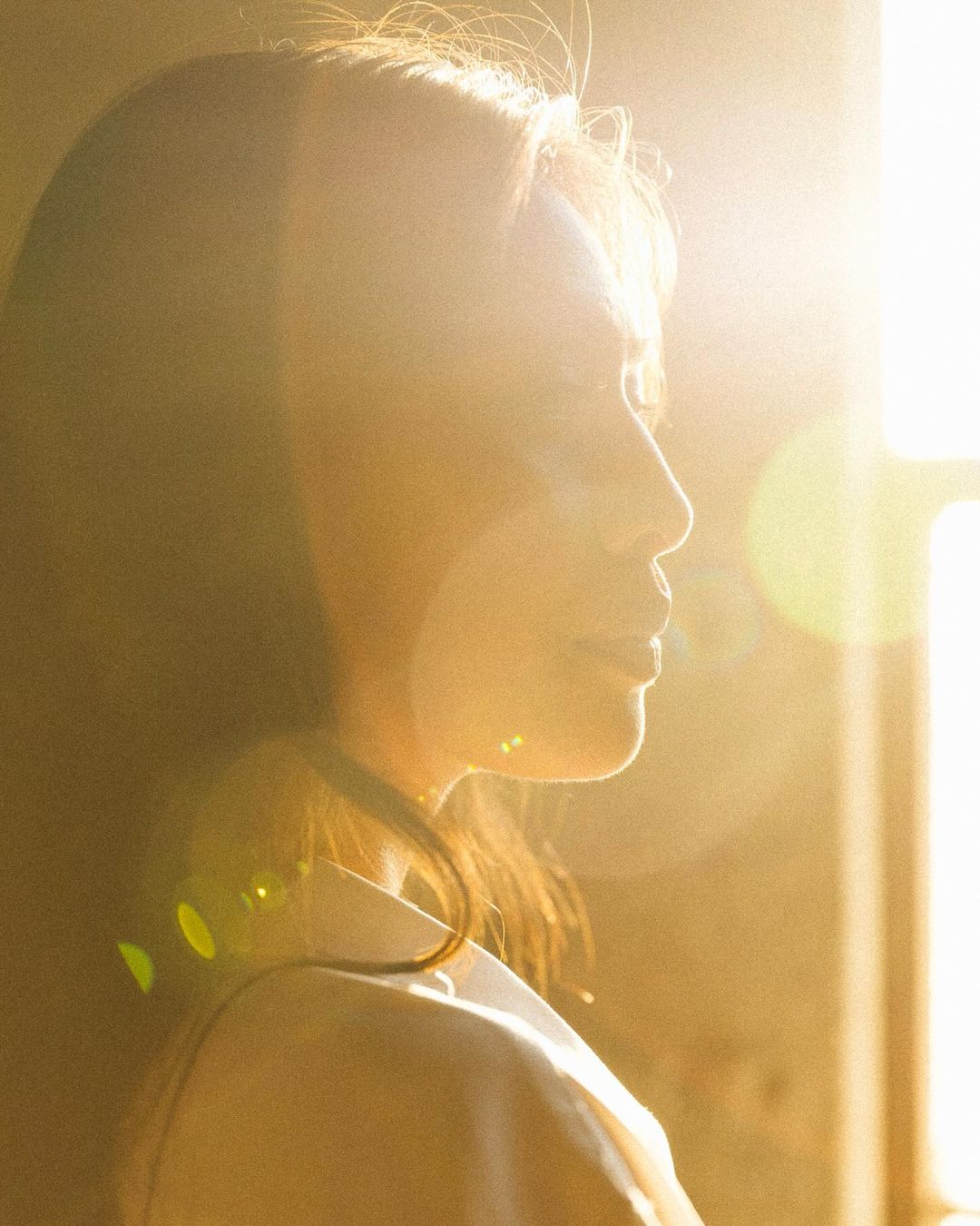 Portrait of a woman in profile near a window, with strong backlighting creating a golden hue. Her hair is long and flowing.