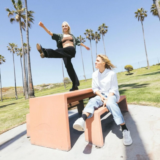 Two young women pose playfully on a concrete bench in a park under a bright blue sky lined with tall palm trees during daytime.