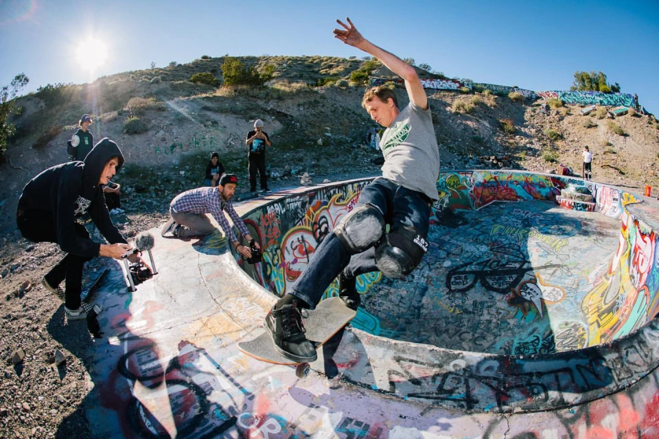 Skateboarder performs a trick in a concrete skatepark bowl, surrounded by other people and graffiti, with a sunny, natural outdoor background.