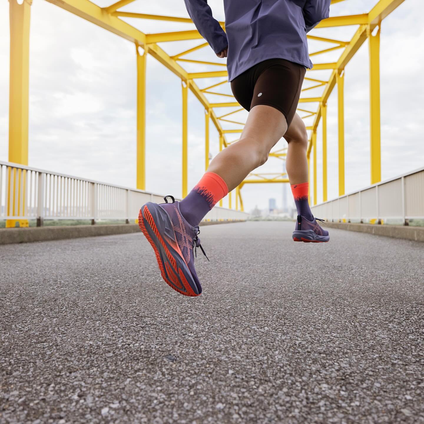 Person running across a yellow bridge wearing athletic shoes, shorts, and socks, with a cityscape visible in the distance.