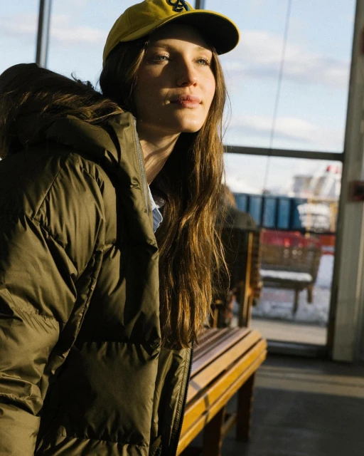 Adult female with long hair poses in a puffer jacket and ball cap while sitting on a bench in an indoor, brightly lit environment.