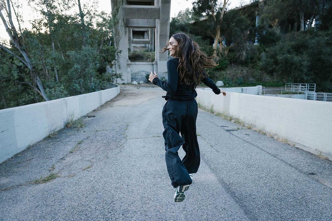 Adult female in black running across an empty road toward a concrete structure surrounded by trees in an outdoor setting during daylight.