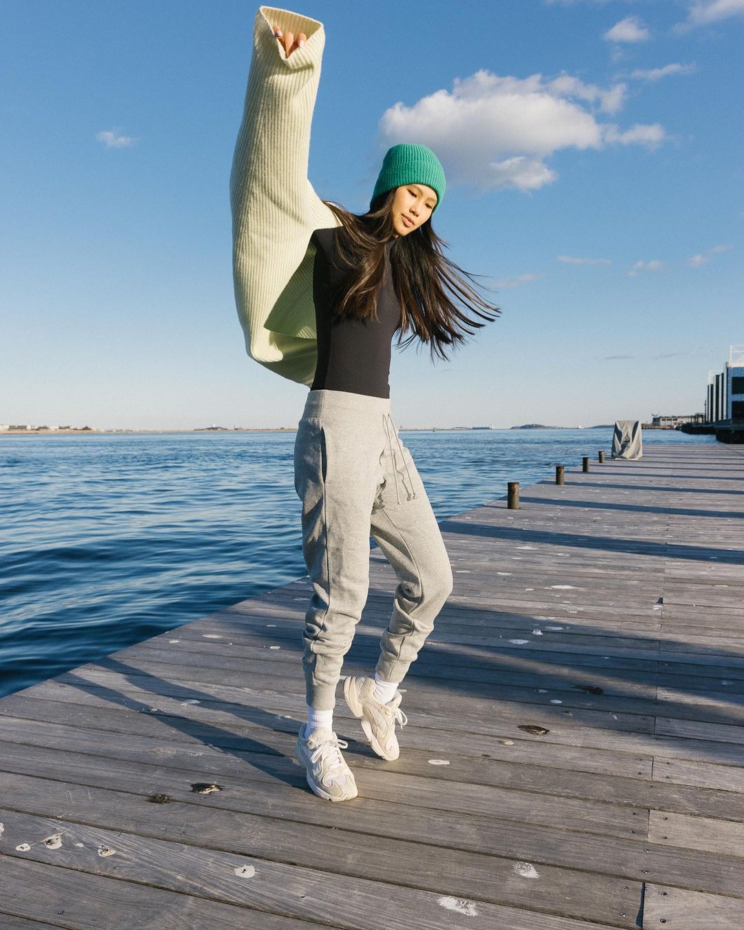 An Asian woman stands on a wooden pier wearing sweatpants and a black shirt, holding a light green sweater and wearing a green beanie.