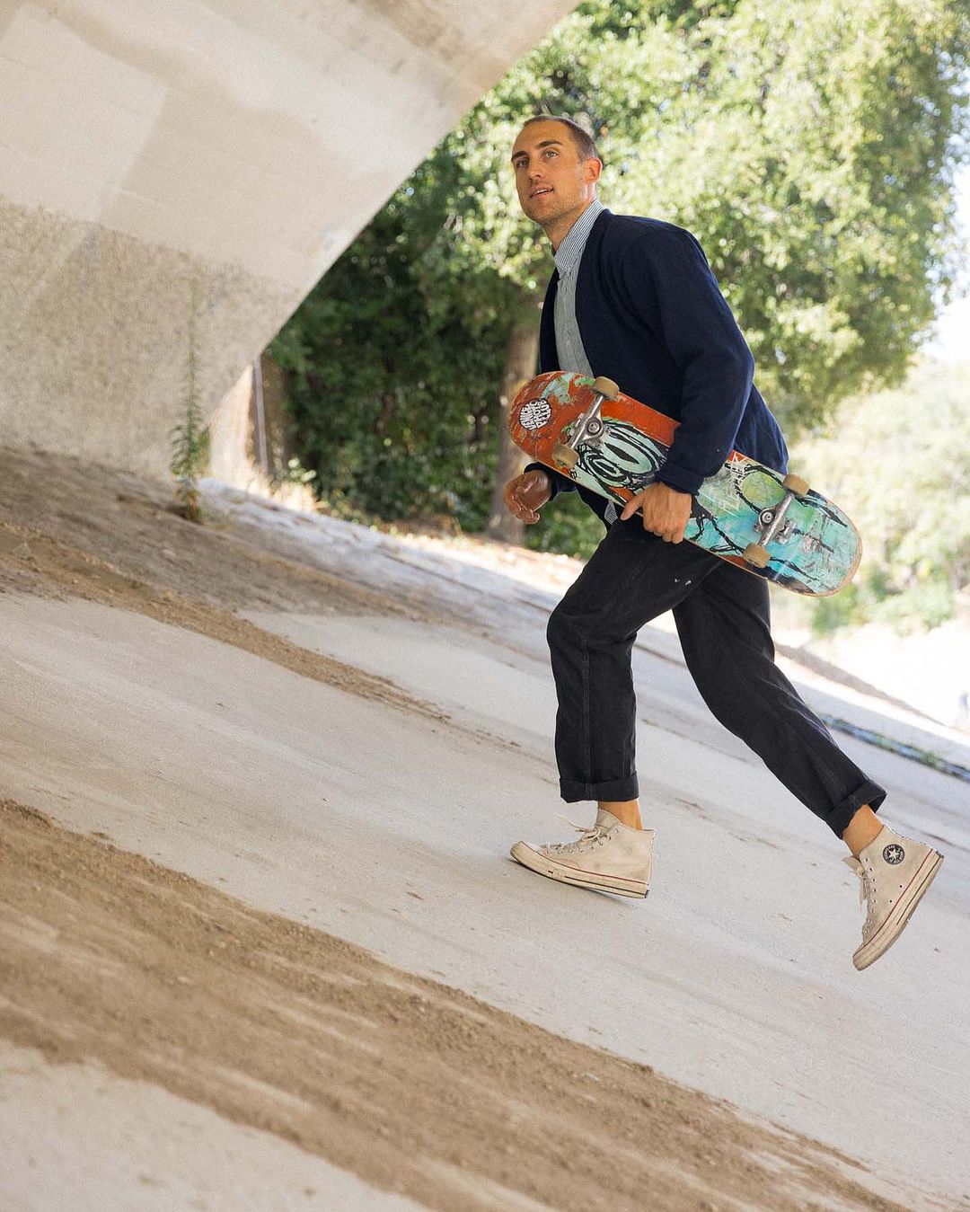 Man in casual attire runs while holding a skateboard outdoors, with a concrete structure and green foliage in the background.