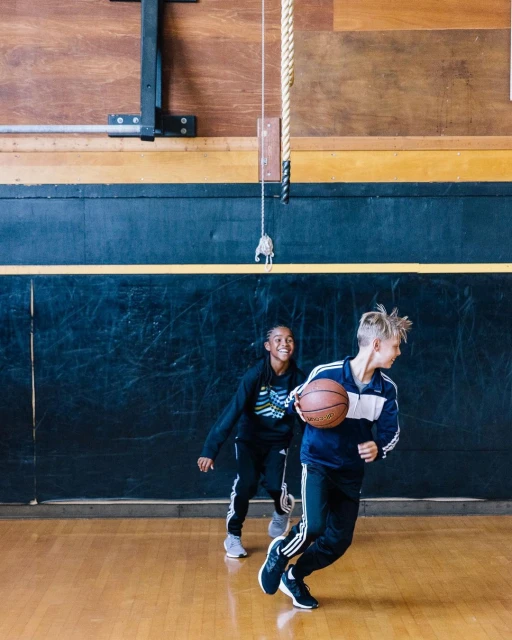 Two young people, a smiling African-American girl and a fair-skinned boy, practice basketball indoors on a polished court with wood paneling.