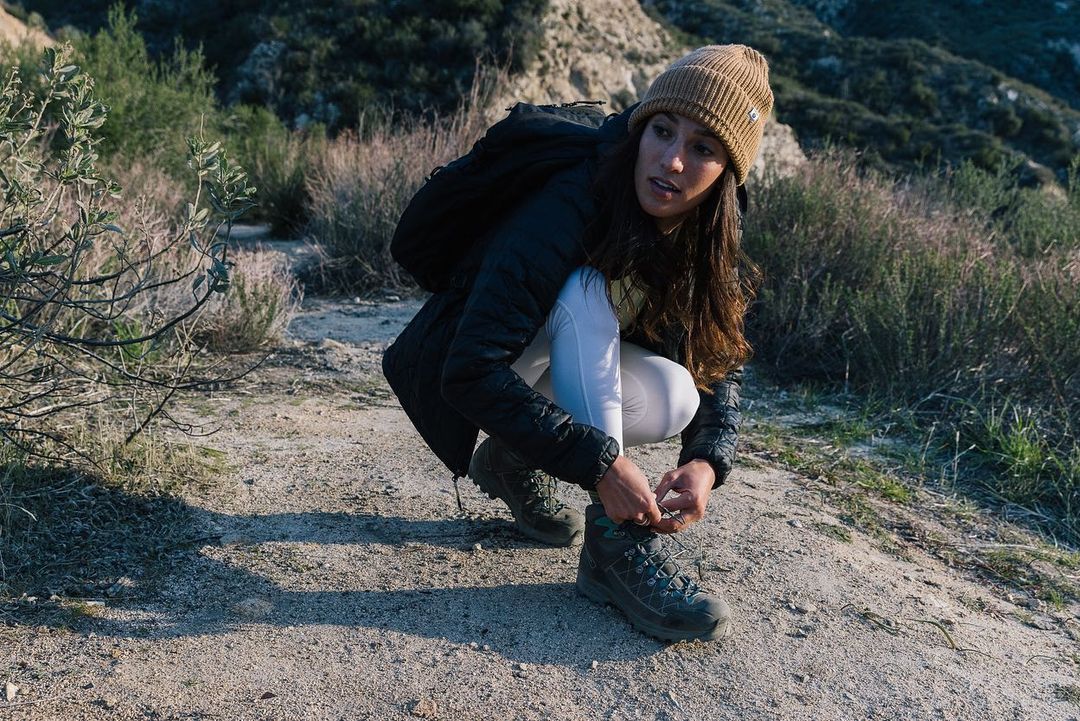 Woman in beanie tying her hiking boot on a dirt path in a sunny outdoor environment with mountains in the background.