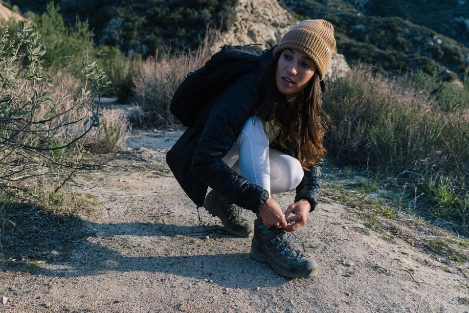 Woman in beanie tying her hiking boot on a dirt path in a sunny outdoor environment with mountains in the background.