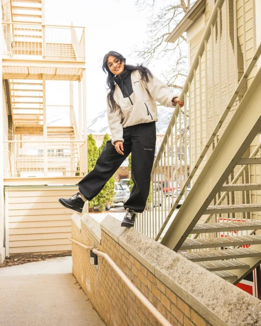 Smiling woman in casual fleece and trousers balances on brick wall in an urban setting, with apartment buildings and mountains in background.