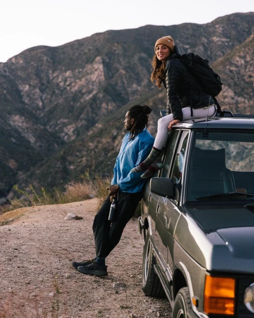 A woman sits on a car roof while a man leans against the car in a mountain setting with a backpack and water bottle at dusk.