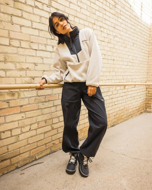 Woman in outdoor clothing poses against a brick wall in natural light, wearing a fleece jacket, black pants, and snow boots.