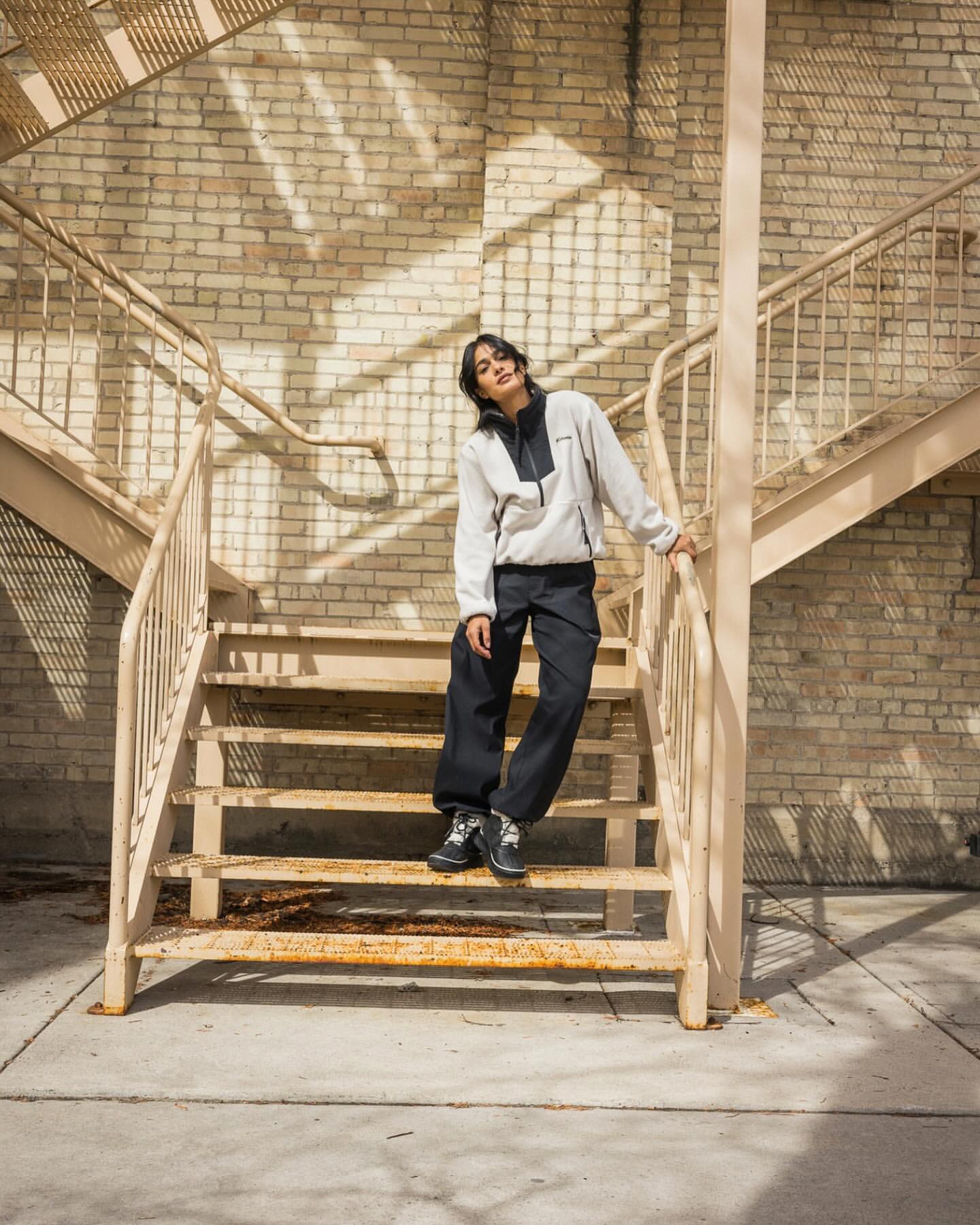 Woman in a fleece jacket and dark pants poses on metal stairs outside in dappled sunlight. Brick wall background and casual fashion style.