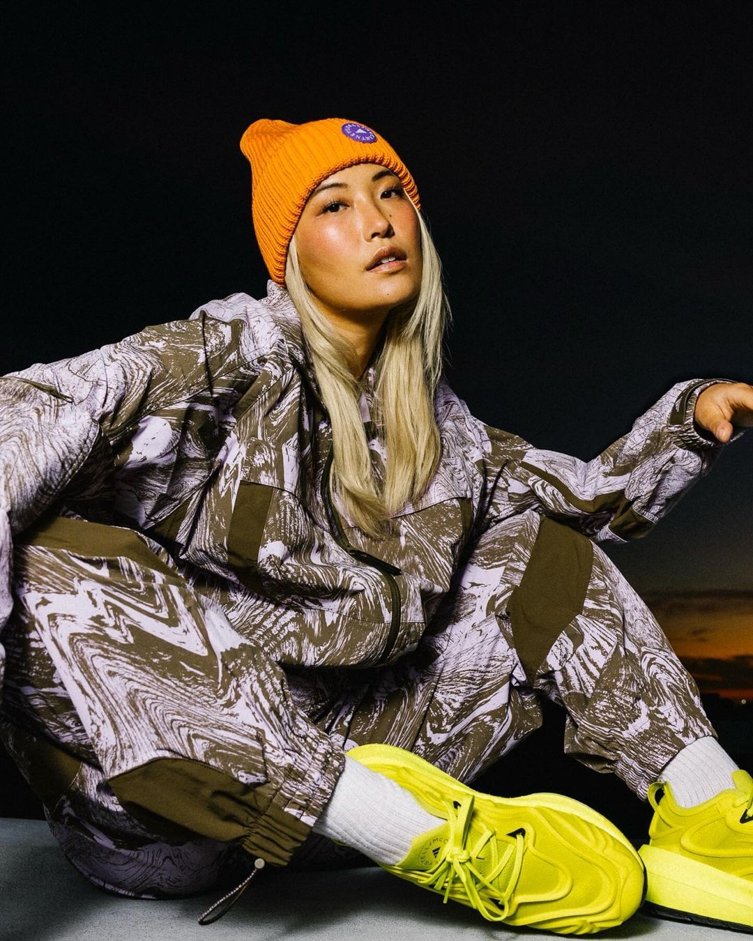 Asian woman in patterned jacket and pants with yellow sneakers, wearing an orange beanie against a dark sky with hints of sunset.