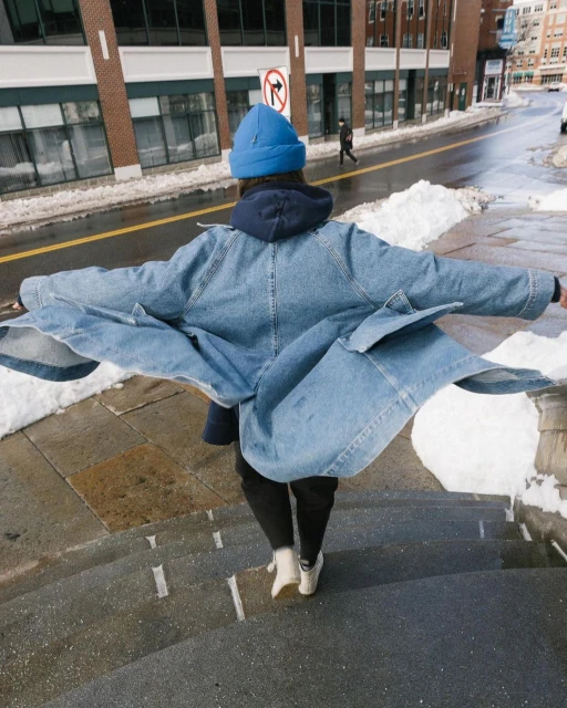 Person in blue denim jacket and beanie walks down stairs on city street in winter with snow, back view, medium shot.