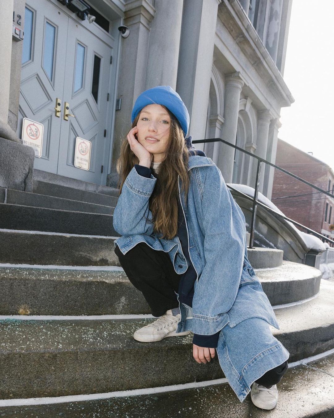 Young woman in blue hat and denim jacket squats on steps of stone building on a bright day, looking at the camera.