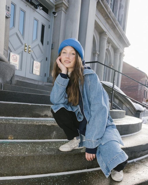 Young woman in blue hat and denim jacket squats on steps of stone building on a bright day, looking at the camera.