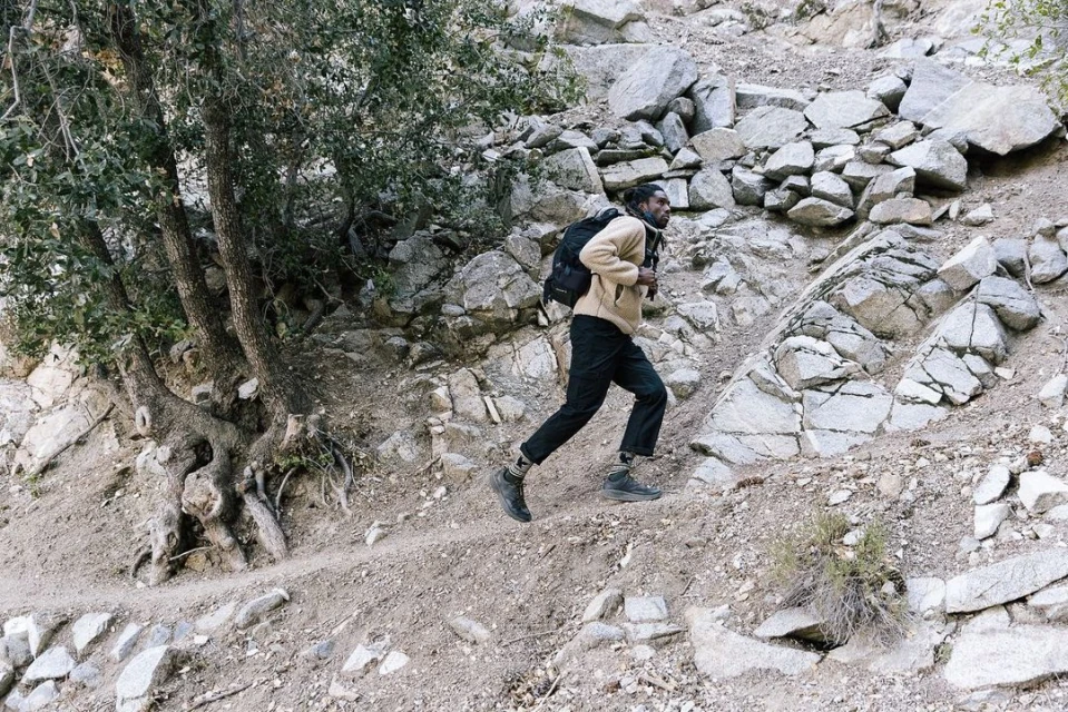 An adult male hiker wearing a tan fleece and black pants ascends a rocky hillside trail with a backpack, surrounded by trees and boulders.