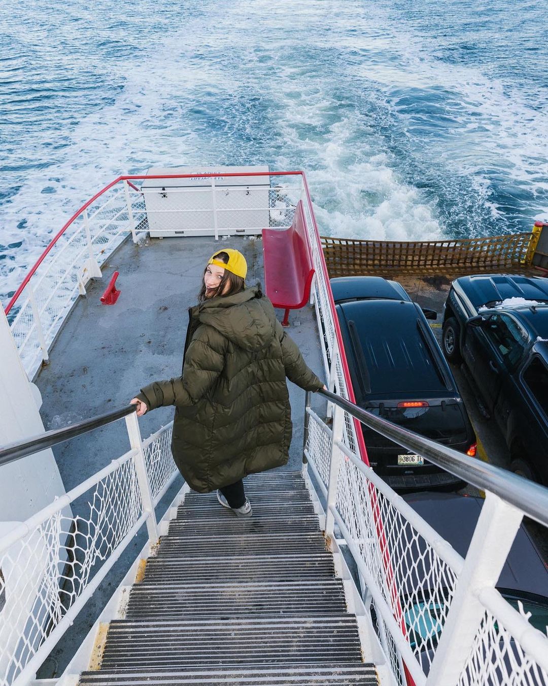 Woman in a puffer coat descends stairs on a ferry, cars parked nearby, the ocean visible in the background with foamy wake.