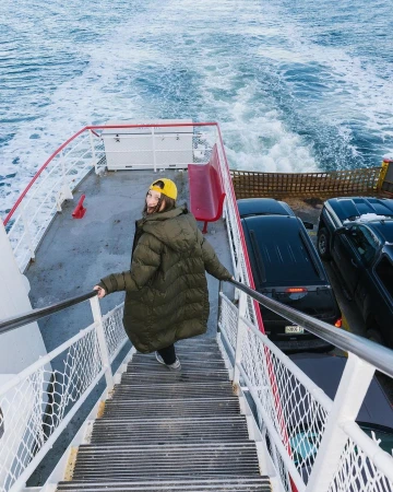 Woman in a puffer coat descends stairs on a ferry, cars parked nearby, the ocean visible in the background with foamy wake.