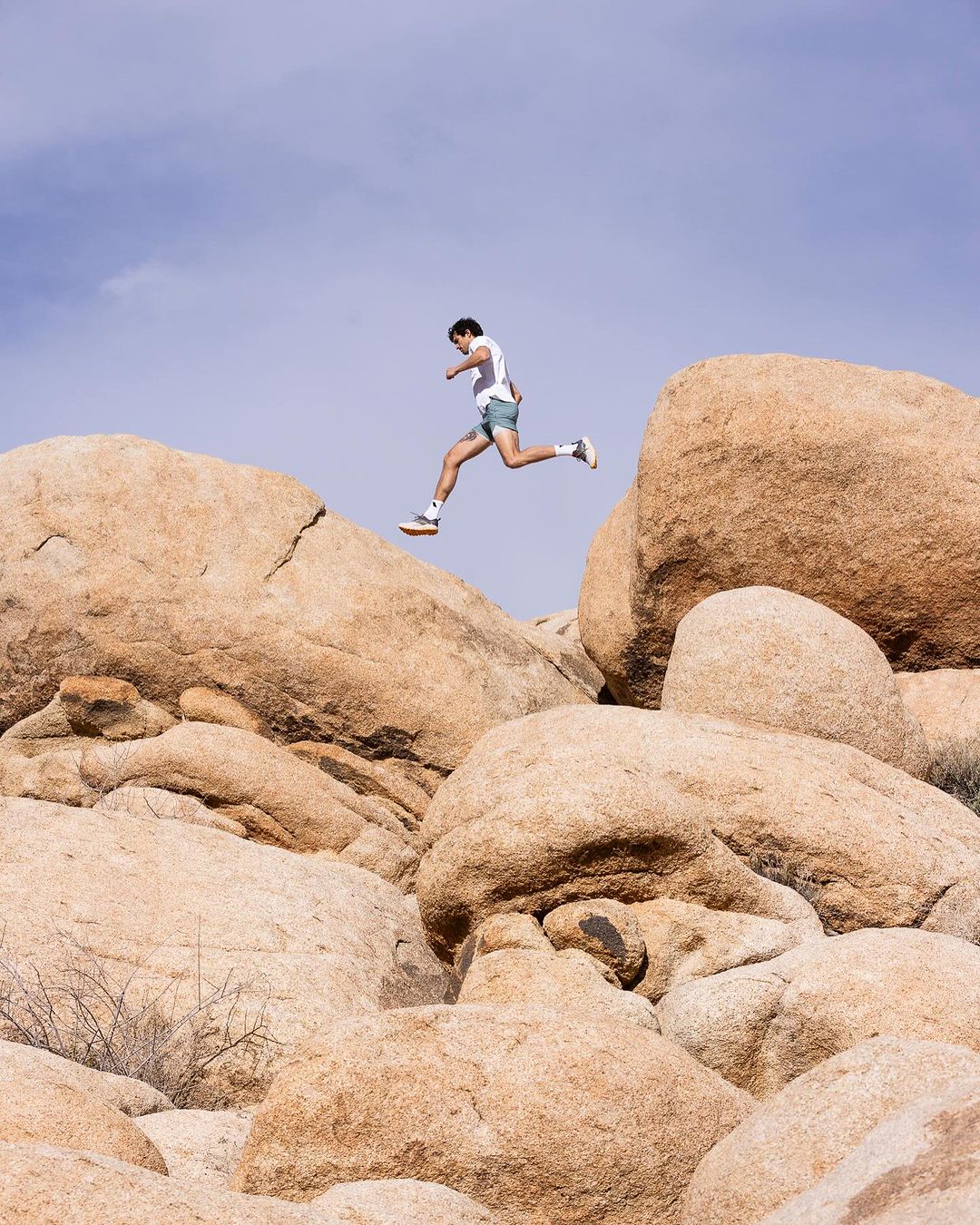 Adult male leaps between tan rocks in a desert setting under a blue sky, wearing athletic shorts and a white t-shirt on a sunny day.