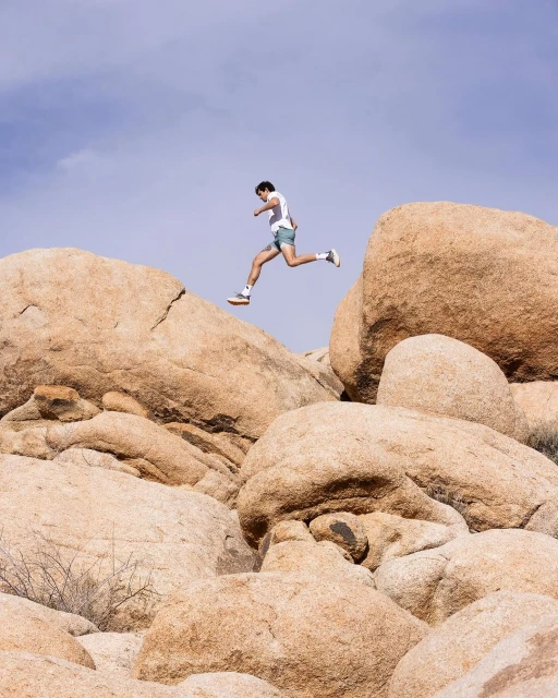 Adult male leaps between tan rocks in a desert setting under a blue sky, wearing athletic shorts and a white t-shirt on a sunny day.