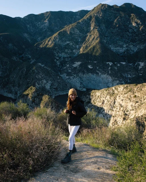 Woman smiles while hiking on a dirt path with mountains in the background on a sunny day with clear skies and green foliage.